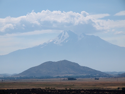 [The image is what appears to be a large shadow mountain superimposed over a darker one in the foreground which is less than half its height. In reality Mount Shasta is just that far in the distance that the haze makes it seem shadowy. The top quarter to a third of Mount Shasta has snow on it. The top peak is in the puffy clouds in the sky.]